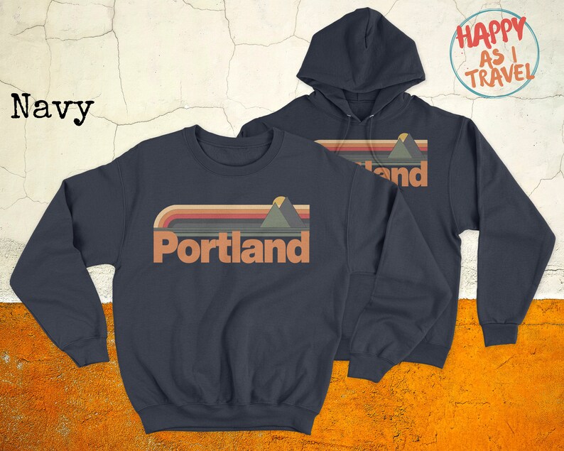May include: Two navy blue sweatshirts featuring a retro mountain and sunset graphic with the text "Portland". One sweatshirt is a crew neck and the other is a hoodie. The hoodie has the text "Happy as I Travel" on the back.
