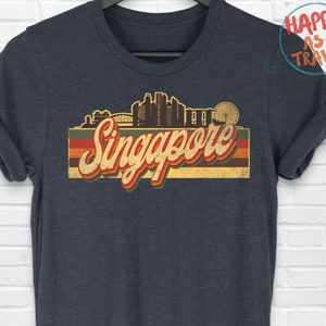 Puede incluir: Camiseta de color gris oscuro jaspeado con un gráfico de estilo vintage del horizonte de Singapur y la palabra "Singapur" en una fuente retro.