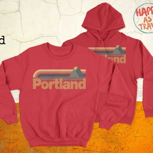 May include: Red sweatshirt and hoodie with a retro-style graphic that says "Portland" with a mountain and sunset design. The text "Happy as I Travel" is on a blue circle in the top right corner.