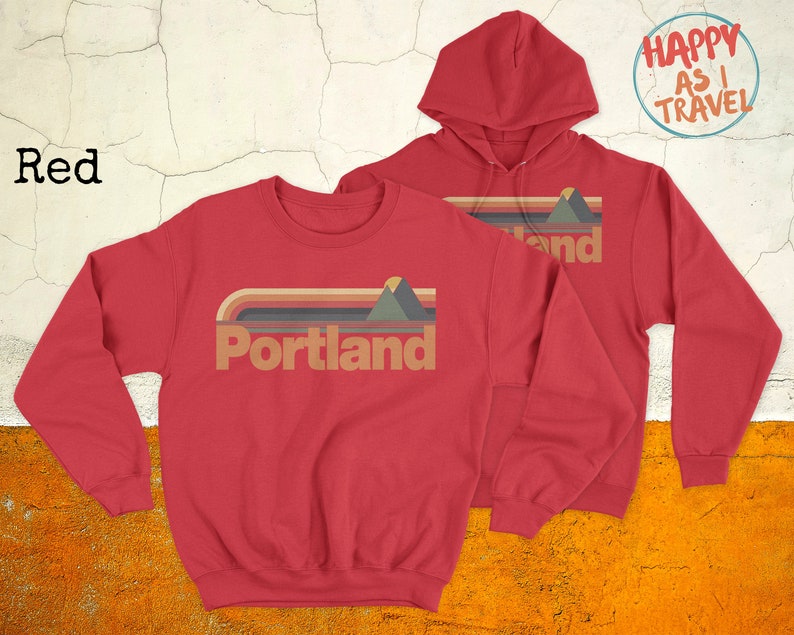 May include: Red sweatshirt and hoodie with a retro-style graphic that says "Portland" with a mountain and sunset design. The text "Happy as I Travel" is on a blue circle in the top right corner.