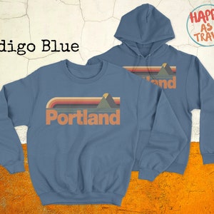 May include: Indigo blue sweatshirt and hoodie with a retro design featuring a mountain range and the word "Portland". The hoodie has a drawstring and the sweatshirt has a crew neck. The text "Happy as I Travel" is printed on a white circle in the top right corner.