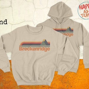 May include: Sand-coloured sweatshirt and hoodie with a retro design. The design features the text "Breckenridge" and a mountain graphic, with horizontal stripes in orange, red, green, and brown. The hoodie has a drawstring hood and the text "Happy as I Travel".