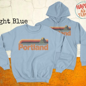 May include: Light blue sweatshirt with a retro-style graphic of mountains and the word "Portland" in a vintage font.