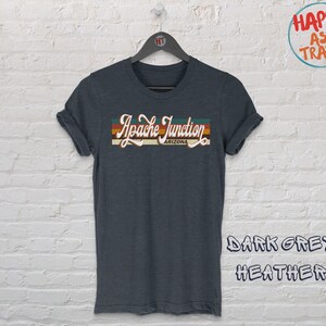 Peut inclure: T-shirt gris foncé chiné avec l'inscription "Apache Junction Arizona" dans un style rétro. Le texte est en blanc, orange, jaune et vert. Le t-shirt est suspendu à un cintre noir.