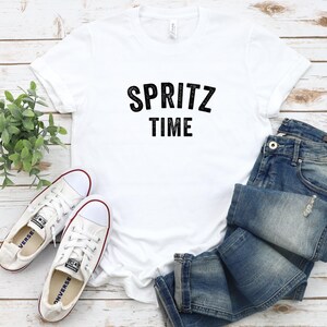 May include: White t-shirt with black text that reads "SPRITZ TIME" on a white wooden background with a pair of white canvas shoes and blue jeans.