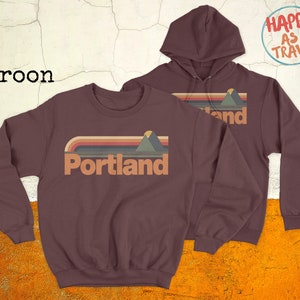 May include: Two maroon sweatshirts featuring a retro-style graphic of a mountain range and the word "Portland". The hoodie has a drawstring and the crewneck has a relaxed fit.