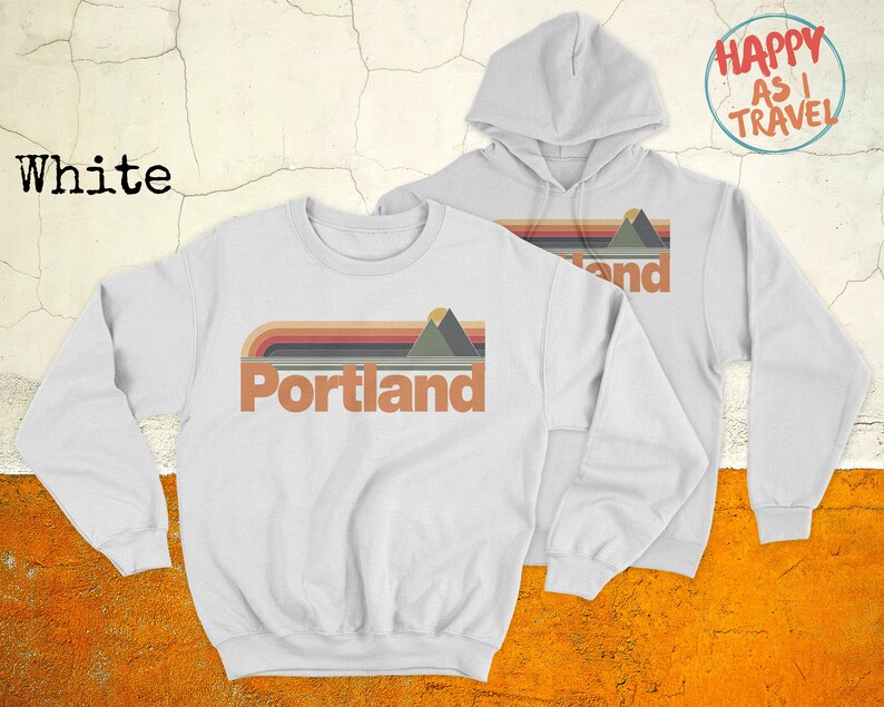 May include: Two white sweatshirts, one with a hoodie. The sweatshirt with the hoodie has the text "Happy as I Travel" in a circle on the upper right side. The sweatshirt without the hoodie has a retro-style graphic with the text "Portland" and a mountain range.
