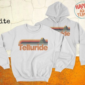 May include: White sweatshirt with a retro mountain graphic and the text "Telluride" in orange. The sweatshirt is shown with a matching white hoodie with the text "Happy as I Travel" on the back.