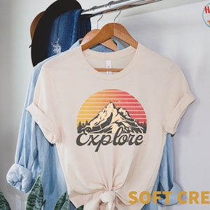 Puede incluir: Una camiseta de color crema con un gráfico de montaña y puesta de sol de estilo vintage. El texto "Explore" está impreso debajo del gráfico. La camiseta está colgada en una percha.