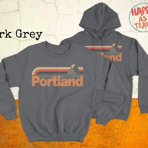 May include: Dark grey sweatshirt with a retro-style graphic of mountains and a sunset, with the word "Portland" in white text. The sweatshirt is shown next to a matching dark grey hoodie with the same graphic and the text "Happy as I Travel" in a circle.