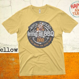 May include: Yellow t-shirt with a graphic of a man with a beard and sunglasses, holding a spatula and a grill. The graphic says "Wood Smoked King of BBQ Dad I only smoke the good stuff".
