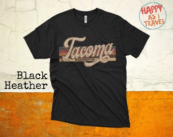 May include: Black heather t-shirt featuring the word "Tacoma" in a vintage font, with "Washington" underneath. The design includes retro horizontal stripes in brown, green, and red. A casual tee.