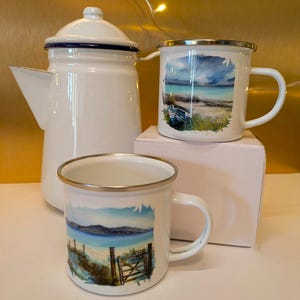 May include: Three white enamel mugs with silver rims. Each mug features a different watercolour painting of a beach scene. The mugs are arranged on a white surface with a white enamel coffee pot in the background.