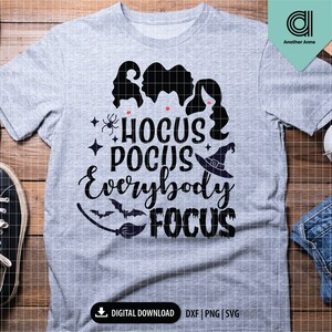 May include: A heather gray t-shirt with the text "Hocus Pocus Everybody Focus" in black. The design includes silhouettes of three witches, a witch hat, a spider, and a bat. The shirt is a digital download.