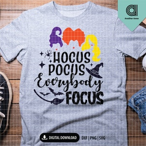 May include: A heather gray t-shirt with the text "Hocus Pocus Everybody Focus" in black script. The design includes silhouettes of witch hair in purple, orange, and yellow, a witch hat, a spider, and a bat. The shirt is a digital download.