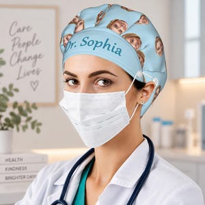 May include: A light blue scrub cap featuring a repeating pattern of a person's face and the text Dr. Sophia printed in dark teal on the front band. The cap includes white buttons on the sides for securing a white face mask and a stethoscope.