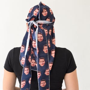 May include: A dark blue head scarf featuring a repeating pattern of a person's face printed across the fabric. The scarf is tied at the back of the head with long, flowing tails that drape downwards, showcasing the consistent facial print design.