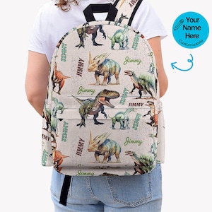 May include: A beige backpack with a repeating dinosaur pattern in green, brown, and orange. The name "Jimmy" is printed in green script. The backpack has black straps and a blue circle with a pencil icon and the text "Your Name Here Customizable".