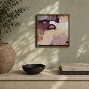 Pu&ograve; includere: Dipinto astratto in cornice dorata, con toni bordeaux, bianchi e beige. A sinistra, un grande vaso marrone strutturato con un ramo frondoso. Una ciotola scura e due libri completano l'esposizione.