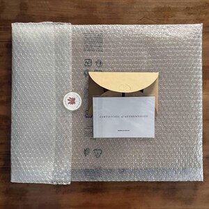 Pu&ograve; includere: Una vista dall'alto mostra un pacco avvolto in pluriball. All'interno, una busta di carta kraft e una carta con la scritta "CERTIFICATE OF AUTHENTICITY" sono visibili. &Egrave; incluso anche un adesivo rotondo con un disegno a foglia.