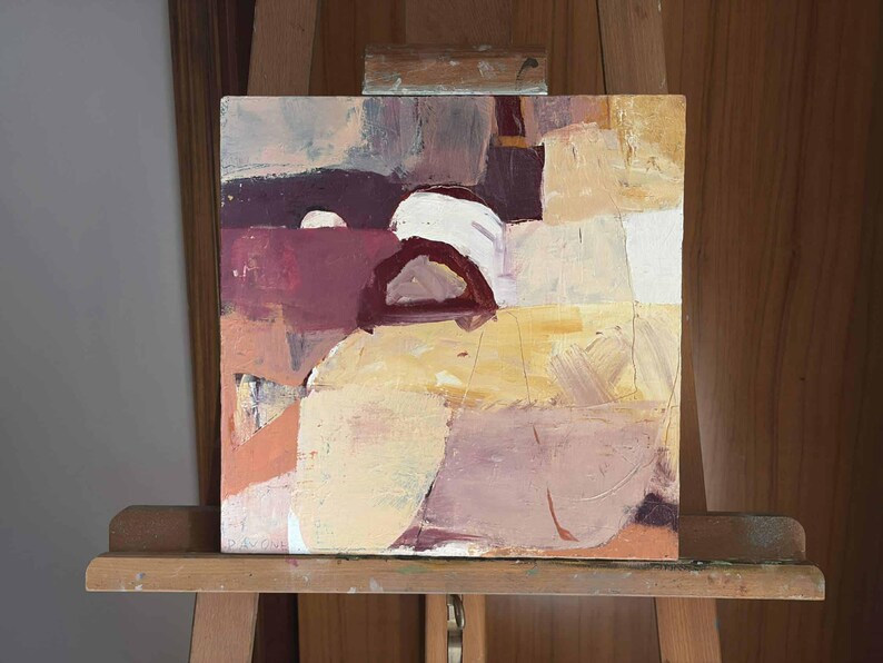 Pu&ograve; includere: Dipinto astratto su tela quadrata, con una tavolozza di colori tenui. L'opera presenta una composizione di forme a strati nei toni del beige, bordeaux e bianco, con pennellate visibili. Il dipinto &egrave; esposto su un cavalletto di legno.