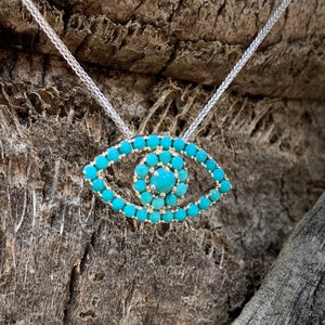 Puede incluir: Un collar de cadena de plata con un colgante de ojo turco turquesa. El colgante est&aacute; hecho de peque&ntilde;as piedras turquesas dispuestas en forma de ojo.
