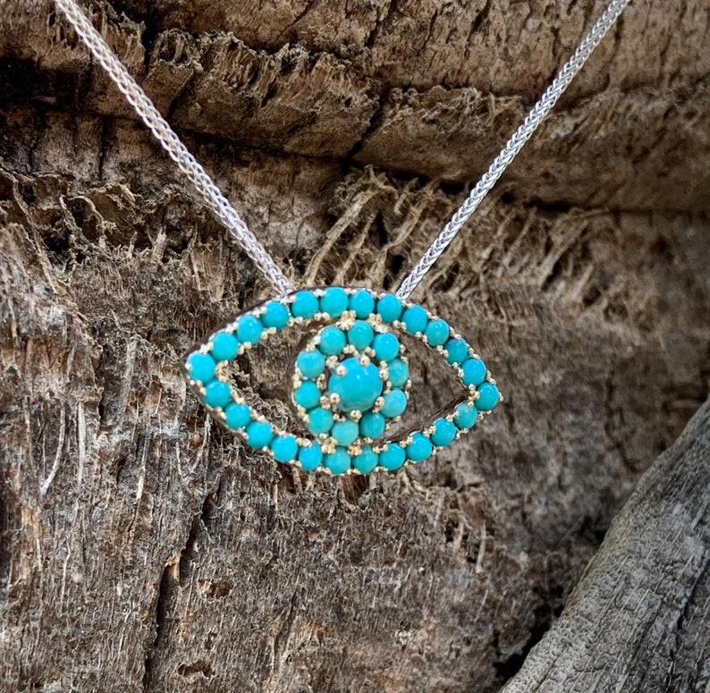 Puede incluir: Un collar de cadena de plata con un colgante de ojo turco turquesa. El colgante est&aacute; hecho de peque&ntilde;as piedras turquesas dispuestas en forma de ojo.