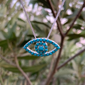 Puede incluir: Un collar de plata con un colgante de ojo turco de color turquesa. El colgante est&aacute; hecho de peque&ntilde;as piedras turquesas dispuestas en forma de ojo.