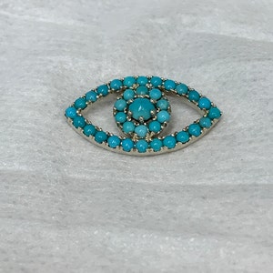 Puede incluir: Un amuleto de ojo de plata con un dise&ntilde;o de ojo turco azul turquesa. El amuleto est&aacute; hecho de peque&ntilde;as piedras redondas turquesas.