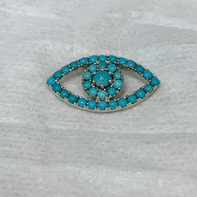 Puede incluir: Un amuleto de ojo de plata con un dise&ntilde;o de ojo turco azul turquesa. El amuleto est&aacute; hecho de peque&ntilde;as piedras redondas turquesas.