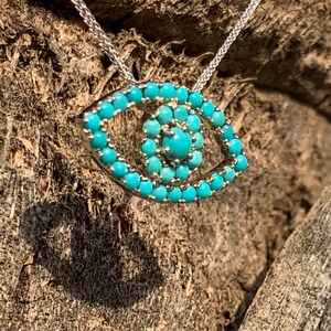Puede incluir: Un collar de cadena de plata con un colgante de ojo turco de color turquesa. El colgante est&aacute; hecho de peque&ntilde;as piedras turquesas engastadas en plata.