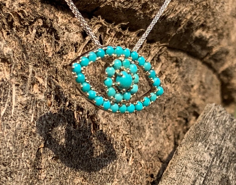 Puede incluir: Un collar de cadena de plata con un colgante de ojo turco de color turquesa. El colgante est&aacute; hecho de peque&ntilde;as piedras turquesas engastadas en plata.