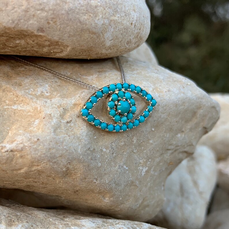 Puede incluir: Un collar de plata con un colgante de ojo turco de color turquesa. El colgante est&aacute; hecho de peque&ntilde;as piedras turquesas dispuestas en forma de ojo.