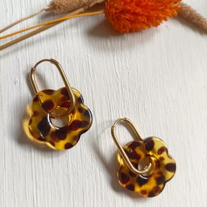 Peut inclure: Deux boucles d'oreilles en forme de fleur avec un motif écaille de tortue marron et jaune. Chaque boucle d'oreille est suspendue à une boucle en métal doré. Elles sont présentées sur une surface blanche, avec des fleurs séchées.