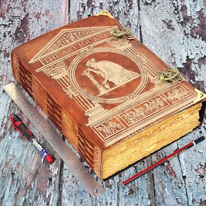 May include: A large, brown leather-bound art journal with gold-colored accents and the title "THE ART JOURNAL" embossed on the cover. A ruler, a red pen, and a pencil are placed next to the journal. The journal measures approximately 12 inches long.