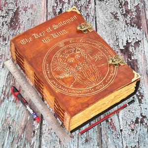 May include: A large, leather-bound book titled "The Key of Solomon the King" with intricate embossed designs and metal clasps. A ruler, pen, and pencil are placed near the book. The book is brown with gold accents.