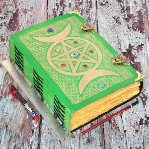 May include: A large, green, leather-bound book with gold accents and a pentagram design on the cover. The book has a clasp closure and is adorned with colorful gemstones. A ruler, pen, and pencil are placed next to the book.