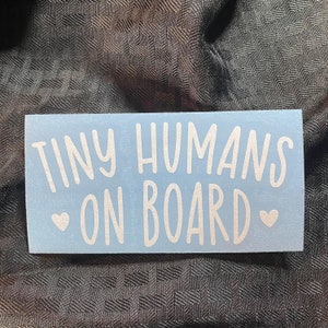 May include: A light blue sticker with the text "Tiny Humans On Board" in white lettering. The sticker has two small white hearts on either side of the text.