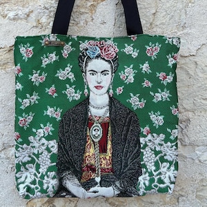 Può includere: Borsa tote verde con un ritratto di Frida Kahlo. La borsa ha manici neri e un motivo floreale bianco e rosso. Il ritratto mostra Kahlo con una corona di fiori e gioielli.