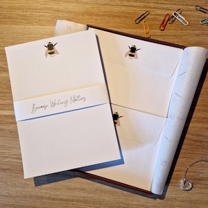 May include: A stationery set with white notecards and envelopes, each decorated with a watercolour bumblebee illustration. The set includes a band with the text "Because Writing Matters". The stationery is in a brown box with paperclips and a necklace in the background.