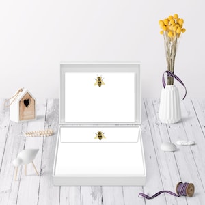 May include: A white box with a white lid, open to reveal a stack of white stationery with a bee illustration on each sheet. The box is on a white wooden surface.