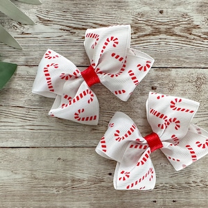 May include: Three white hair bows with a red candy cane pattern and a red center. The bows are arranged on a light wood grain surface, suggesting a holiday or Christmas theme.