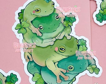 May include: A sticker featuring a stack of three green frogs with closed eyes, surrounded by shamrocks. The frogs are stacked on top of each other, with the largest frog at the bottom and the smallest frog at the top. The sticker has a white border and the text "Bubbles Art Craft" is visible in the background.