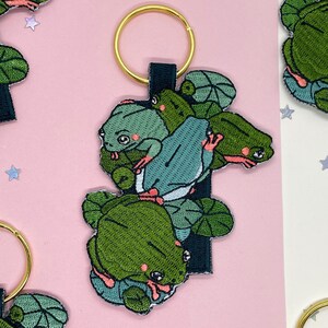 Puede incluir: Llavero bordado de rana con un grupo de ranas verdes con detalles rosas y una rana azul claro en el centro. El llavero tiene un anillo dorado y un respaldo de tela negra. El fondo es rosa.