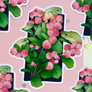 Puede incluir: Patr&oacute;n repetido de pegatinas con ranas verdes posadas en ramas con bayas rosas y hojas verdes. Las pegatinas est&aacute;n colocadas sobre un fondo rosa con acentos en forma de estrella dispersos.