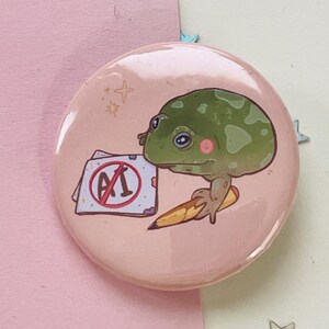 May include: A pink button with a green frog holding a pencil and a piece of paper with a red circle and the letters "A1" crossed out.