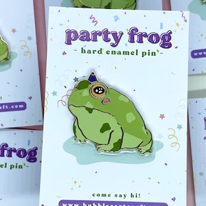 Puede incluir: Un pin de esmalte de rana verde de fiesta con un gorro de fiesta morado y un contorno dorado. La rana lleva un gorro de fiesta y tiene un rubor rosa en las mejillas. El pin está en una tarjeta blanca con el texto "party frog - hard enamel pin" y "come say hi! www.bubblesartcraft.com".