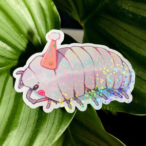 Può includere: Un adesivo olografico di un cartone animato di un porcellino di terra con un cappello da festa. Il porcellino di terra è viola chiaro con glitter iridescenti e un viso sorridente. Il cappello da festa è rosa con pois arancioni. L'adesivo è su uno sfondo di foglie verdi.