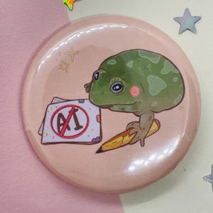 Może przedstawiać: Różowy guzik z zieloną żabą trzymającą ołówek i kartkę papieru z czerwonym kółkiem i przekreślonym tekstem "A1".