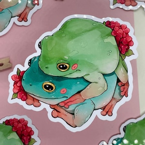 Puede incluir: Dos ranas verdes de árbol con flores rojas en la espalda, una rana está encima de la otra, la rana de arriba es verde y la rana de abajo es azul.